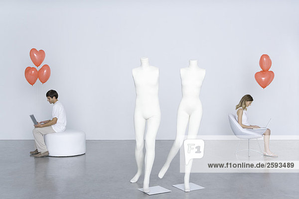 Man and woman sitting with laptops and heart balloons  mannequins in foreground