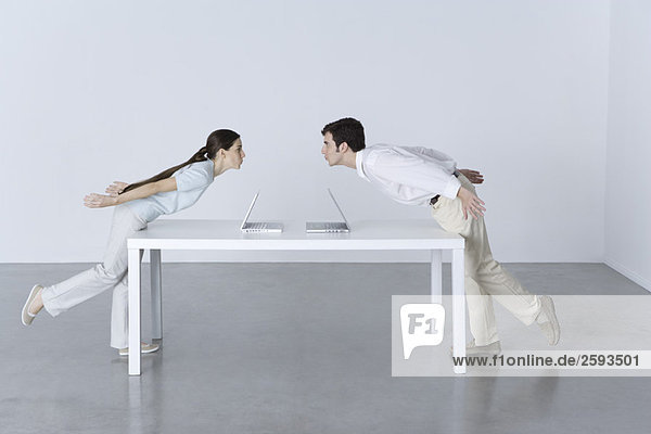 Mann und Frau an gegenüberliegenden Enden des Tisches  die sich aufeinander zubeugen  Laptops zwischen ihnen