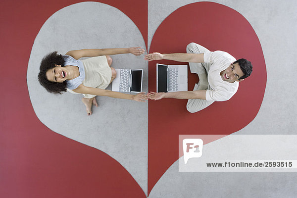 Man and woman sitting with laptops on large heart  arms out  touching hands  overhead view