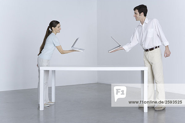 Man and woman at opposite ends of table  holding laptop computers toward one another