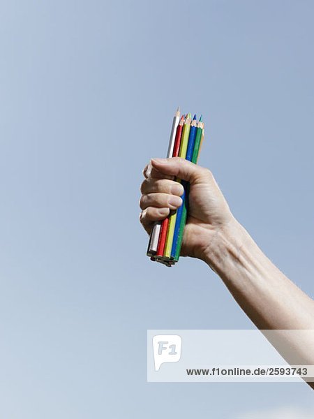 Eine menschliche Hand hält eine Handvoll Buntstifte gegen einen blauen Himmel.