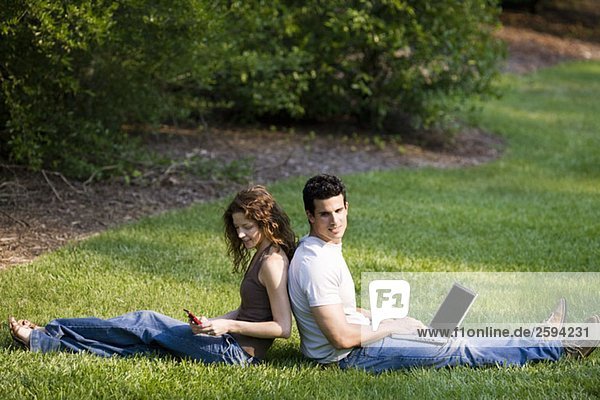 Ein junges Paar sitzt Rücken an Rücken im Gras mit drahtlosen Geräten.