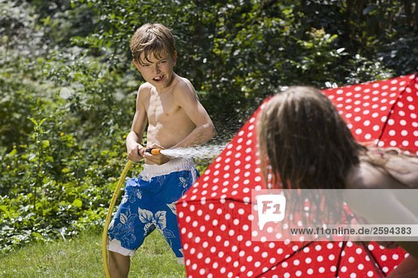 Ein kleiner Junge  der seine Schwester mit einem Gartenschlauch besprüht.