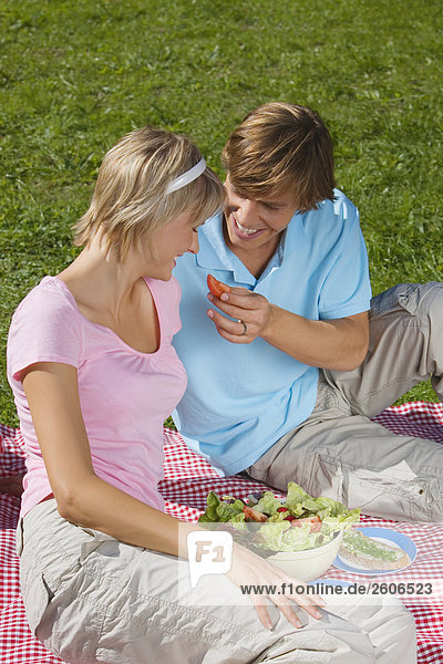 Junges Paar im Park beim Picknick mit Salat Mann gibt Frau ein Stück Tomate