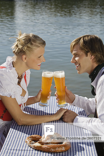 Junges Paar in Tracht sitzt handhaltend am Biertisch mit Breze prosten sich mit Weizenbier zu Biergarten am Seehaus Englischer Garten