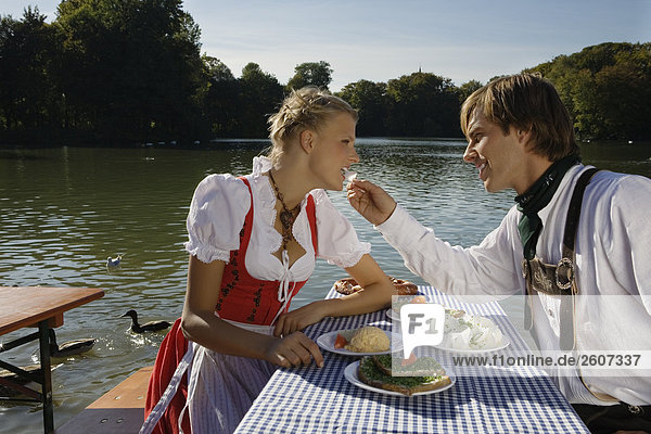 Junges Paar in Tracht sitzt am Biertisch mit Essen Mann füttert Frau mit Radi Biergarten am Seehaus Englischer Garten