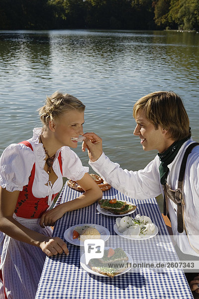 Junges Paar in Tracht sitzt am Biertisch mit Essen Mann füttert Frau mit Radi Biergarten am Seehaus Englischer Garten
