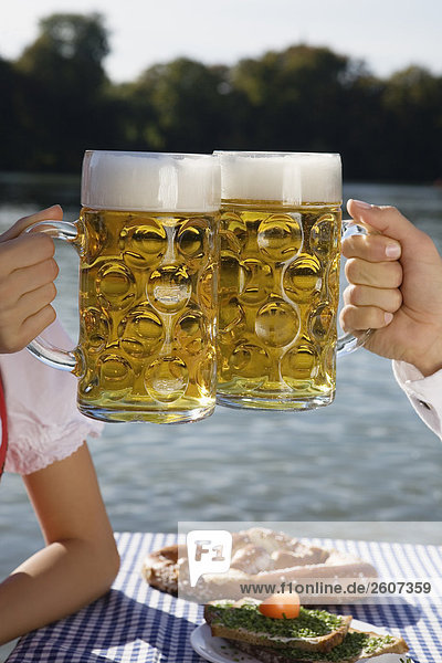 Junges Paar in Tracht sitzt am Biertisch prosten sich mit einer Mass Bier zu Biergarten am Seehaus Englischer Garten Nahaufnahme der Masskrüge