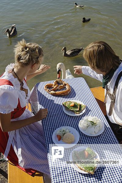 Junges Paar in Tracht sitzt am Biertisch mit Essen schauen auf Schwan im See Biergarten am Seehaus Englischer Garten