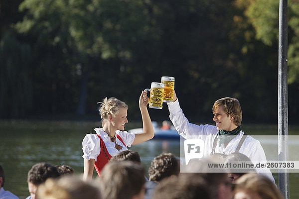 Junges Paar in Tracht steht im Biergarten am Seehaus Englischer Garten prosten sich mit einer Mass Bier zu Menschenmenge im Vordergrund