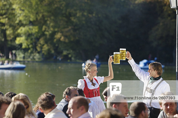 Junges Paar in Tracht steht im Biergarten am Seehaus Englischer Garten prosten sich mit einer Mass Bier zu Menschenmenge im Vordergrund