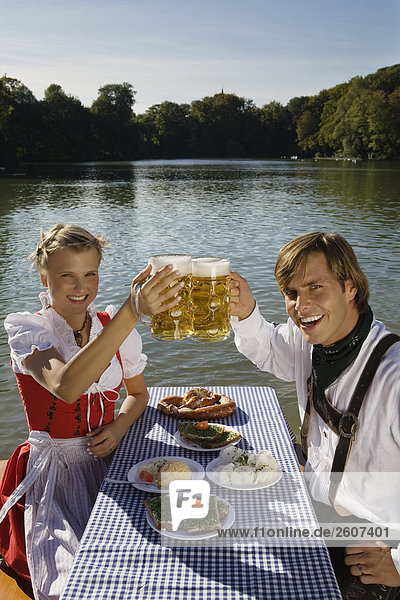 Junges Paar in Tracht sitzt am Biertisch mit Essen prosten sich mit einer Mass Bier zu im Biergarten am Seehaus Englischer Garten