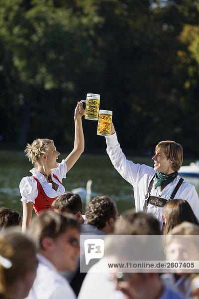 Junges Paar in Tracht steht im Biergarten am Seehaus Englischer Garten prosten sich mit einer Mass Bier zu Menschenmenge im Vordergrund