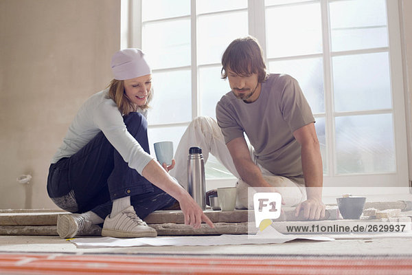 Young couple sitting on floor of empty room  examining plans