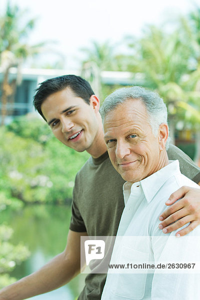 Young man with arm around father's shoulder  both smiling  portrait