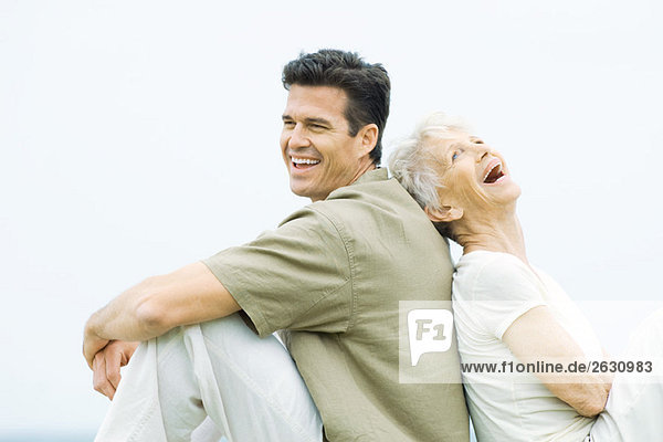 Seniorin und erwachsener Sohn sitzen Rücken an Rücken  lachend  Seitenansicht