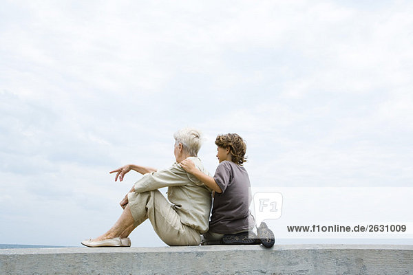 Großmutter und Enkel sitzen zusammen auf einer niedrigen Wand im Freien und schauen auf die Aussicht.