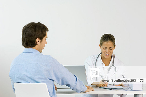 Ärztin sitzt gegenüber dem Patienten am Schreibtisch und schreibt Notizen auf