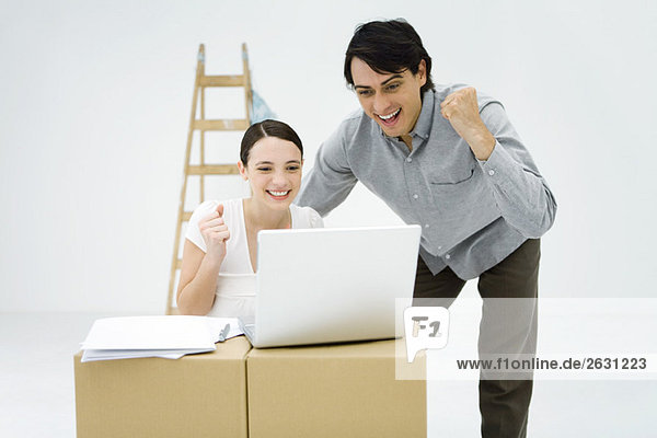 Junges Paar schaut auf Laptop-Computer auf Karton  lächelnd  geballte Fäuste