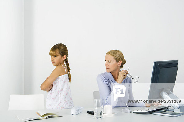 Frau sitzt vor dem Computer und schaut über die Schulter auf die Tochter  die in der Nähe schmollt.