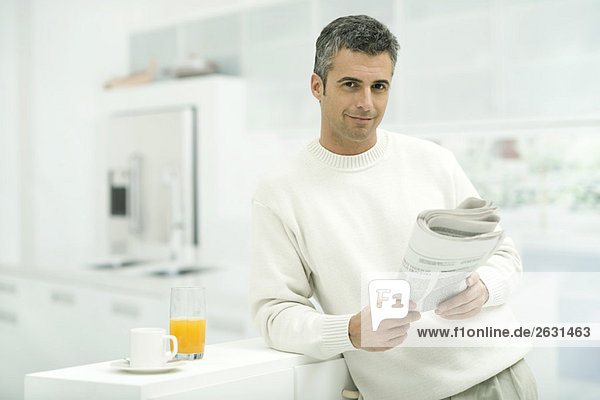 Man leaning against kitchen counter  holding newspaper  smiling at camera