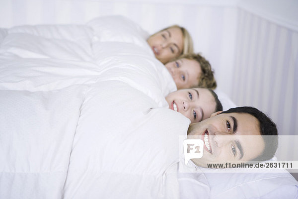 Familie liegt zusammen im Bett unter der Bettdecke und lächelt in die Kamera.