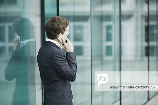 Junger Geschäftsmann steht vor Glasschiebetüren und telefoniert mit dem Handy