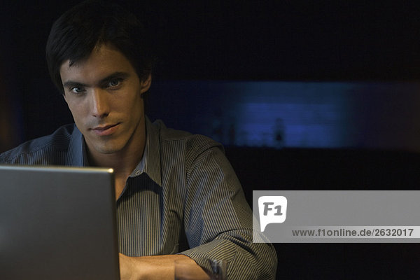 Mann im dunkel erleuchteten Raum am Tisch mit Laptop  Blick auf Kamera  Porträt