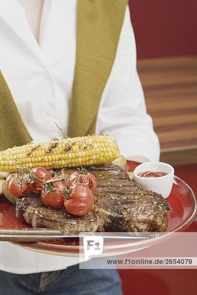 Frau hält T-Bone-Steak mit Maiskolben und Kirschtomaten