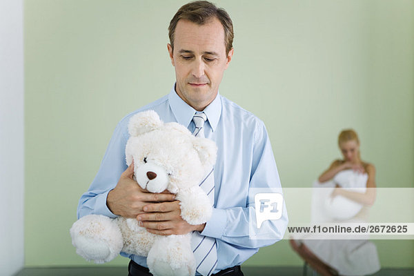New father holding teddy bear  looking down  wife holding baby in background