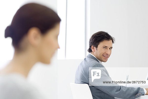 Professional man sitting at desk  smiling over shoulder at camera  female colleague in foreground