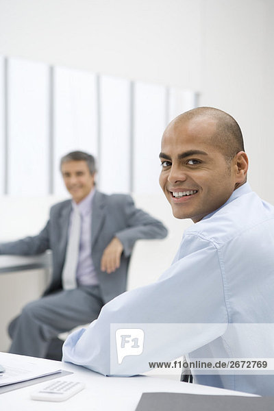 Businessman sitting at desk  smiling over shoulder at camera  colleague in background