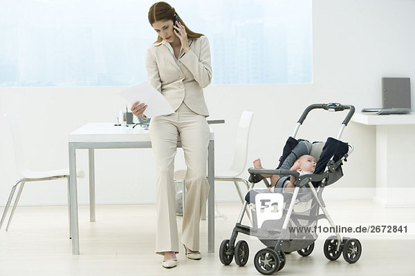 Professionelle Frau im Büro mit Baby im Kinderwagen  im Gespräch auf dem Handy
