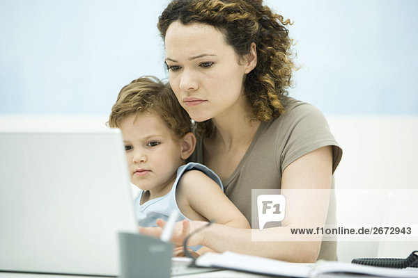 Mutter hält Kleinkind-Sohn im Schoß  während sie den Laptop benutzt.