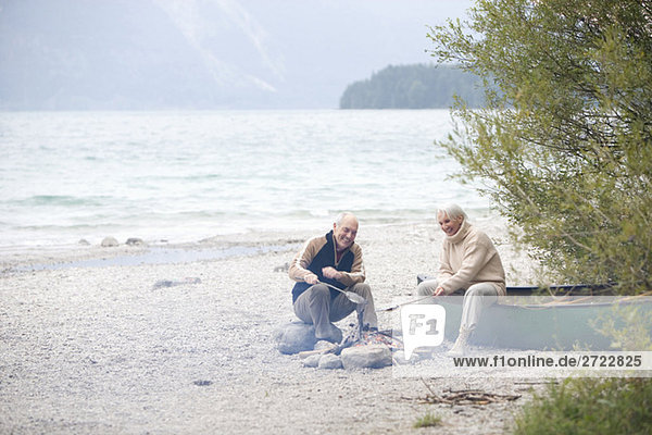 Germany, Bavaria, Walchensee, Senior couple sitting at campfire, grilling fish