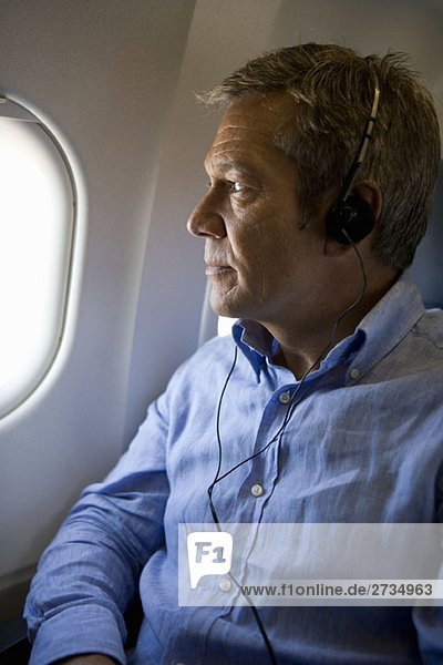 Ein männlicher Passagier  der Kopfhörer hört und aus dem Fenster eines Flugzeugs schaut.