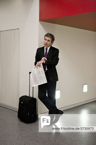Ein Geschäftsmann  der mit seinem Koffer wartet und eine Zeitung hält.