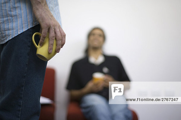 Man casually holding coffee cup at side  colleague sitting in background