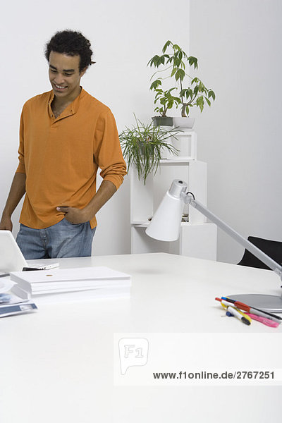 Casual man standing by desk with hand in pocket  looking down at laptop