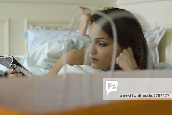 Young woman lying in bed reading magazine  shopping bag in foreground