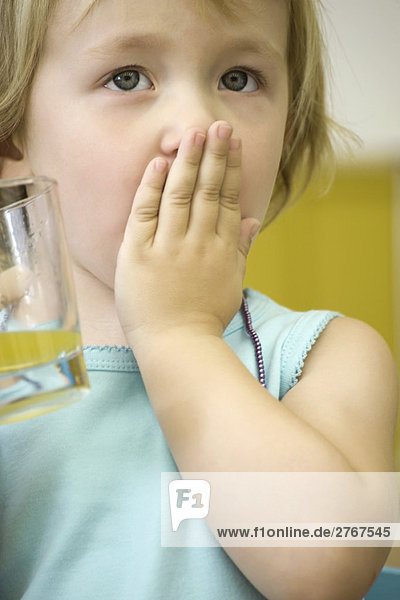 Kleines Mädchen hält ein Glas Saft und bedeckt den Mund mit einer Hand.