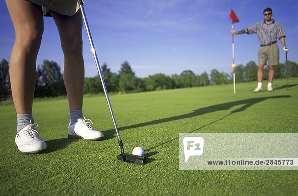 Weibliche Golfer Putts auf Golf grün wie männliche Golfer hilft durch Anheben der Flagge von der Loch  British Columbia  Kanada.