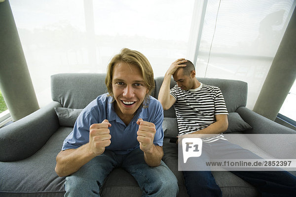 Zwei Männer  die auf dem Sofa sitzen und fernsehen  einer jubelt  der andere hält den Kopf.