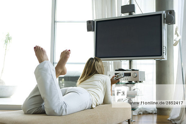 Frau auf dem Bauch liegend  Flachbildfernseher mit Surround Sound  Fernbedienung in der Hand  Rückansicht