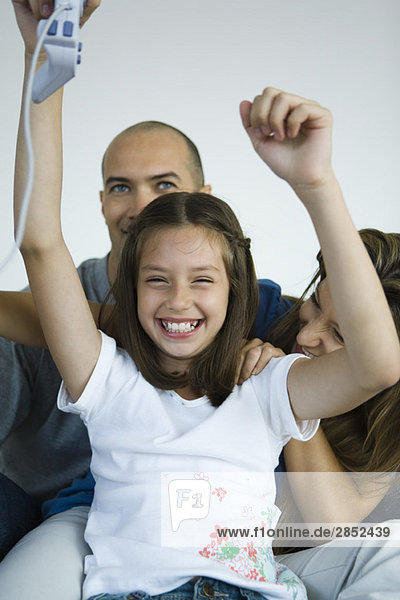 Mädchen sitzt mit Eltern  hält Videospiel-Controller in der Luft und lächelt