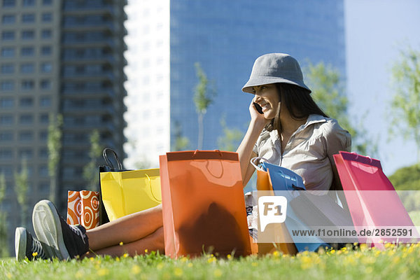 Junge Frau auf dem Boden sitzend  umgeben von Einkaufstaschen  am Handy sprechend