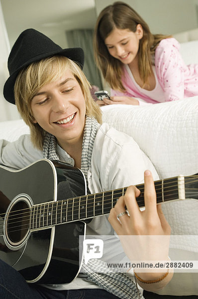 Teenager-Junge spielt Gitarre und seine Schwester mit dem Handy hinter ihm.