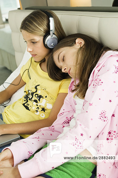 Girl working on a laptop and listening to headphones and her sister sleeping on her shoulders