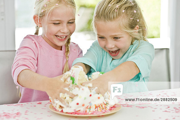 Zwei Mädchen  die spielerisch ihre Hände in einen Geburtstagskuchen stecken.