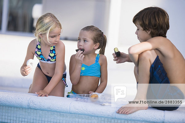 Zwei Mädchen und ein Junge essen Schokoladen-Donuts am Pool.